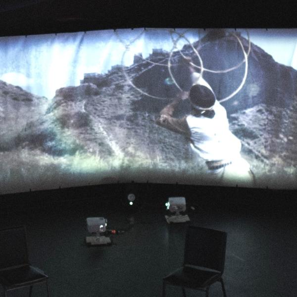 A person hoop dances on a panoramic screen in a large circular space.