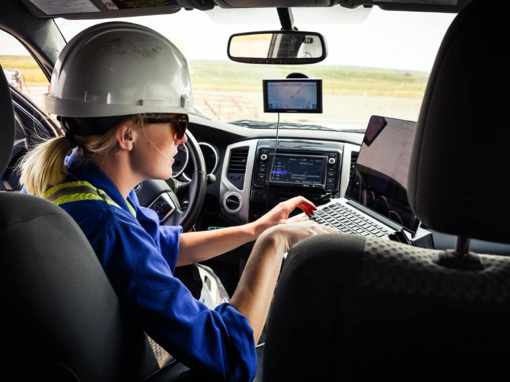 A young researcher wearing a hardhat sits in the driver’s seat of a vehicle, typing on a laptop.