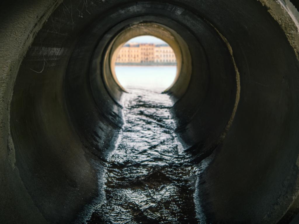 Dark liquid flows through a concrete pipe with a river and cityscape visible through the end of the pipe