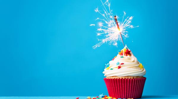 A photo of a vanilla cupcake in a red cupcake holder with a lit sparkler in it, in front of a baby blue background.
