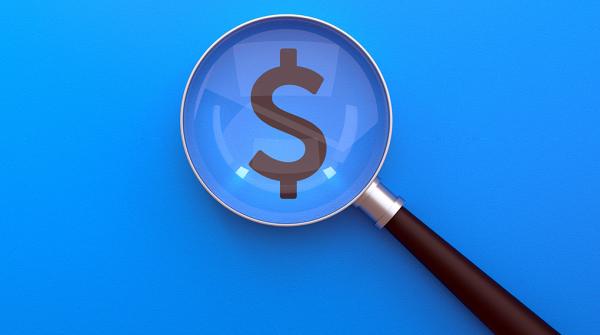 A stock image of a hand-held magnifying glass that is magnifying a bold, black dollar symbol printed on a bright blue background. 