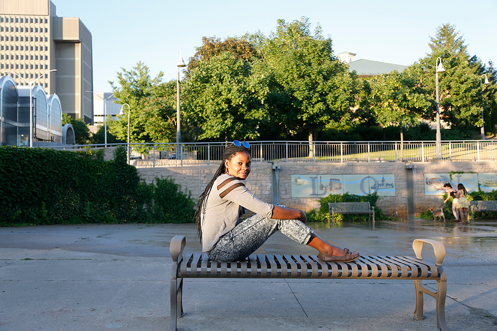 Jacqueline Mboko, an interpreter for London's Cross Cultural Learner Centre, holding a red binder posing for a photo on a park bench.