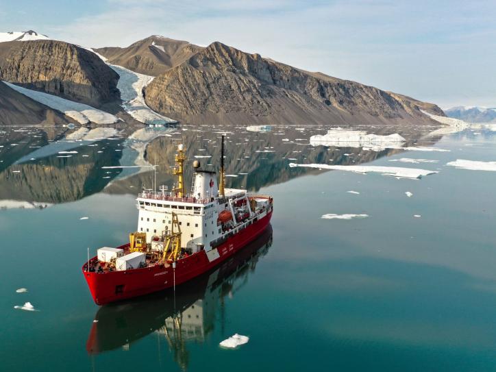 A large red and white icebreaker glides through deep blue water, surrounded by big chunks of floating ice.