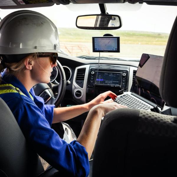 A young researcher wearing a hardhat sits in the driver’s seat of a vehicle, typing on a laptop.
