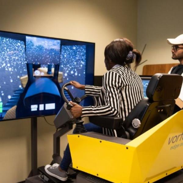 Vue de dos d’une personne assise aux commandes d’un simulateur dont elle tient le volant sous le regard d’une autre personne à sa droite qui porte une casquette blanche.