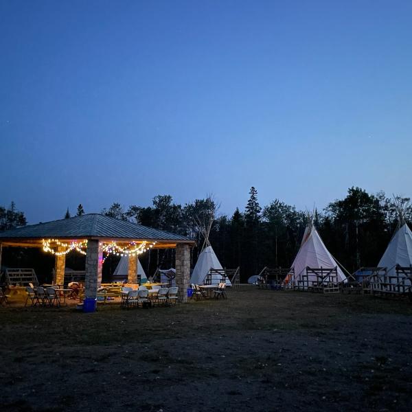 An outdoor scene at dusk with a stone pavilion and several tipis arranged in a circle with bleachers.