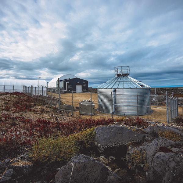 A rocky outcrop with low brush in the foreground with a silver cylindrical building in the middle ground behind a fence and a large domed structure in the background