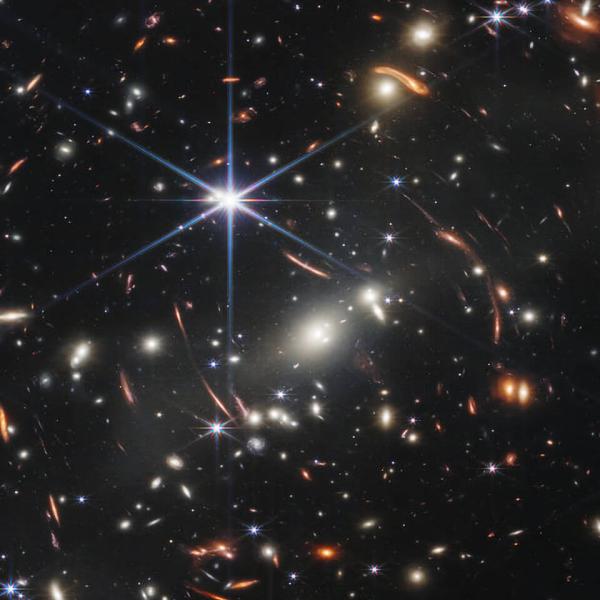 Distant galaxies appear as bright glowing spots in this Webb telescope image, with some smeared by gravitational lensing; foreground stars appear bright with six-pointed diffraction spikes, owing to the shape of Webb's mirrors. 
