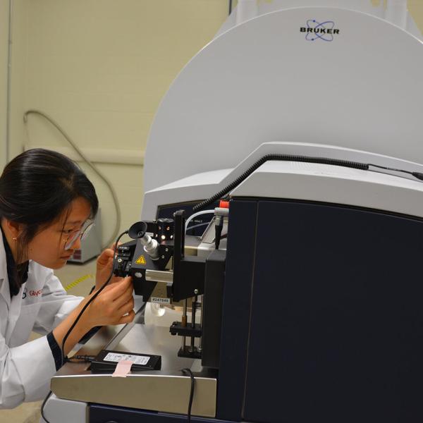 Une personne en sarrau blanc observe attentivement un instrument rattaché à un gros appareil noir qui ressemble à un scanner dans un laboratoire.