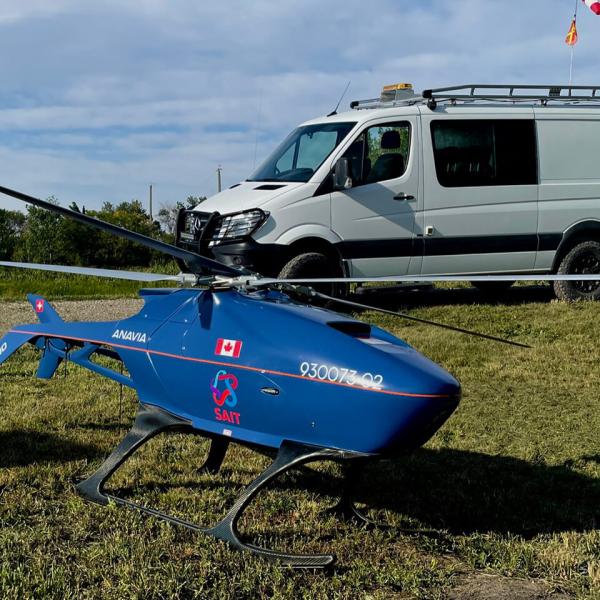 A drone resembling a small blue helicopter sits on a grassy area in the foreground with a white van parked on a driveway in the background.