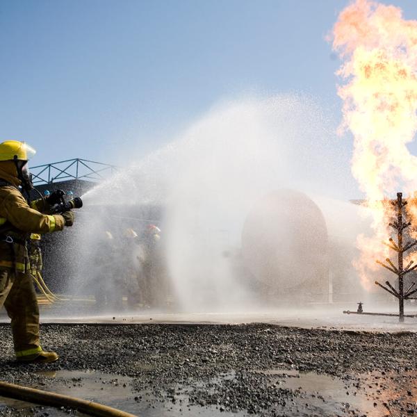 Photo of a fire simulation exercise. Two people wearing fire coats aim a fire hose at a burning building. 