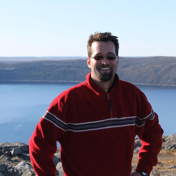 Guillaume St-Onge is wearing a striped red sweater and sunglasses, posing in front of the Pingualuit crater in Quebec, full of clear blue water.
