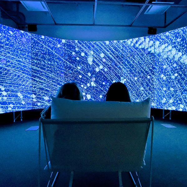 Two people sit in front of a curved video wall displaying illuminated renderings of satellites orbiting earth.