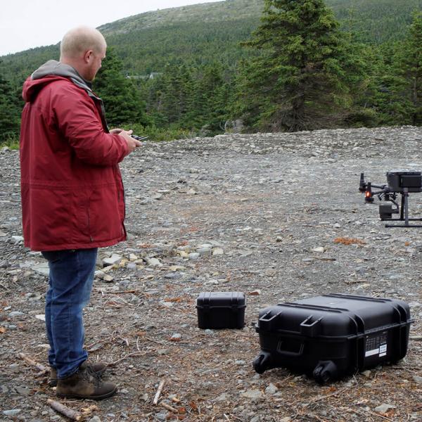 A man in a red jacket holds the remote control as a drone takes off from a rubble-strewn site. 