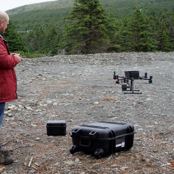 Un homme vêtu d’un manteau rouge tient une télécommande alors qu’un drone décolle d’un site jonché de débris.