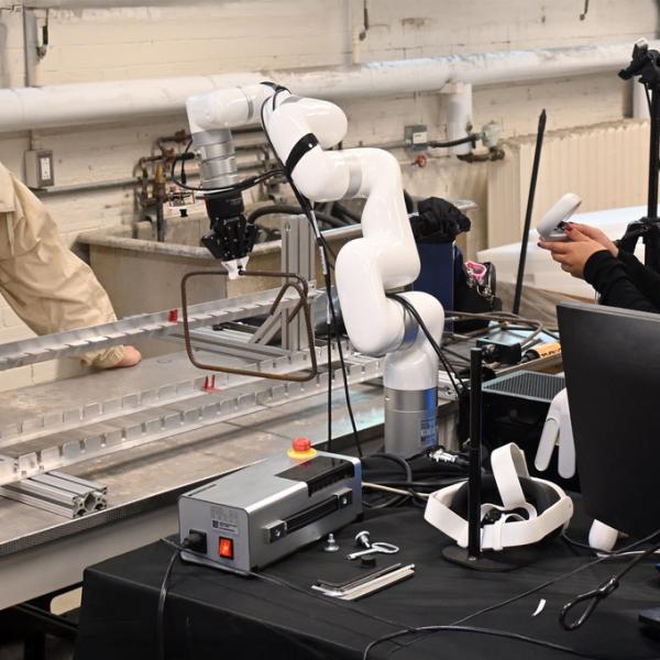 A multi-hinged robotic arm holds a rectangle formed from rebar. On the right, a young woman holds a small white box in her hands. On the left, a young man looks at the frame into which the rebar is being inserted.
