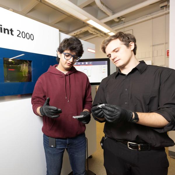 Two students wearing disposable gloves each hold a small metal object as they stand in front of a large TRUMPF TruPrint 2000 machine.