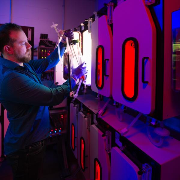 A scientist inside a lab pulls a large, clear cylinder from inside one of several illuminated chambers.