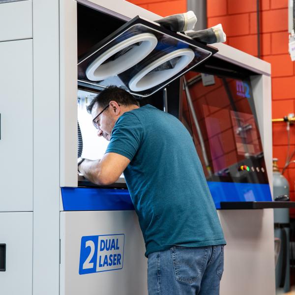 A man leans into the building chamber of a large 3D printer. A control panel is to his right. 