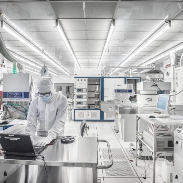 A researcher in hooded white cleanroom coveralls, mask and gloves types on a laptop in a white-tiled cleanroom packed with high-tech equipment.