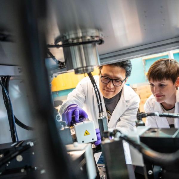 Two scientists in lab coats and gloves examine the contents of a metal chamber equipped with high-tech components.
