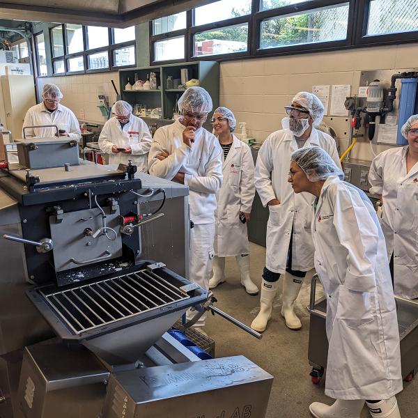 Seven people wearing boots, disposable hairnets, and white lab coats, along with protective goggles, are gathered around a MilkyLab machine in a laboratory located in a basement bathed in light from a row of windows at the very top and all along the wall. Some of them are looking at their cell phones, others are smiling, and still others have approached the cheese-making machine and seem to be wondering how it works.