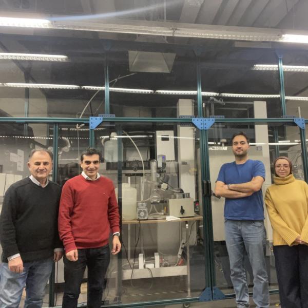Four researchers stand in front of a large, glassed-off section of a lab filled with experimental equipment.