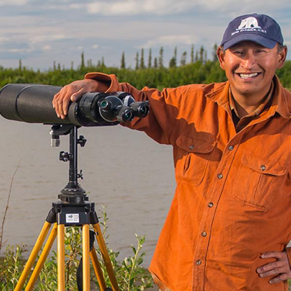 A man stands beside a river with his arm resting on a set of long-range observation binoculars.