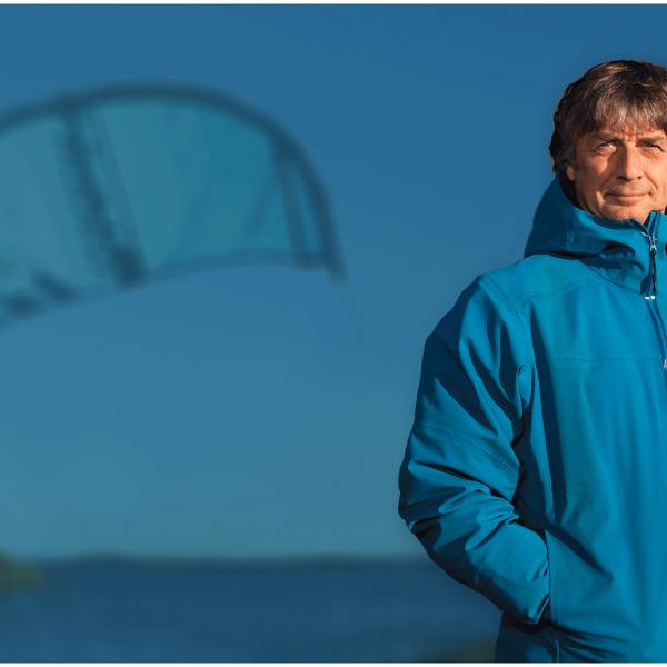 Richard Myerscough poses in the foreground with a large kite from a kiteboard in the sky in the background.