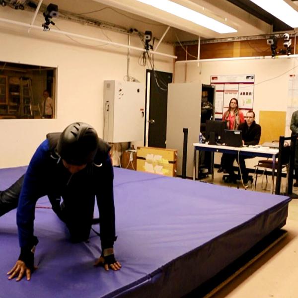 Young man falling on a large purple mattress, wearing a helmet and protective pads, while researchers in the back of the falling laboratory monitor the impact of his fall on a computer.