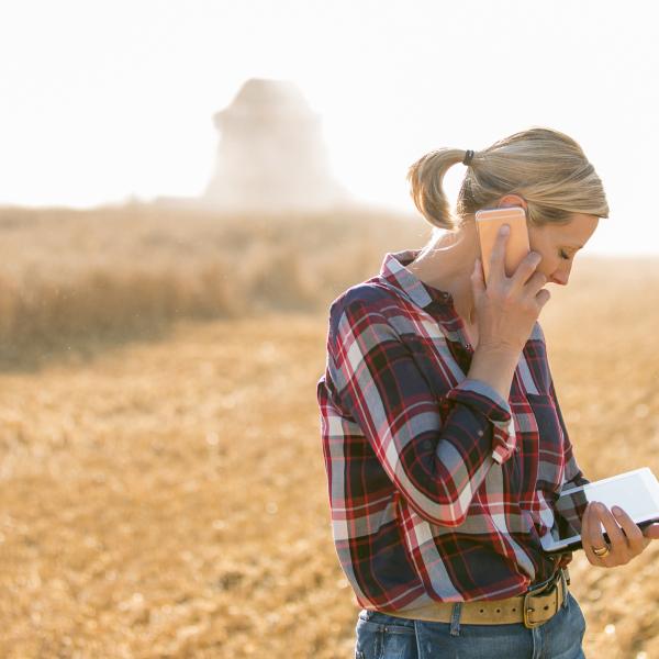 A woman stands in a fallow field talking on a smart phone and consulting a tablet.
