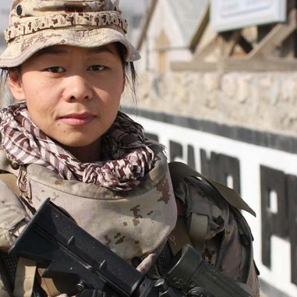 A woman in a military uniform faces the camera, angling a machine gun at the ground.