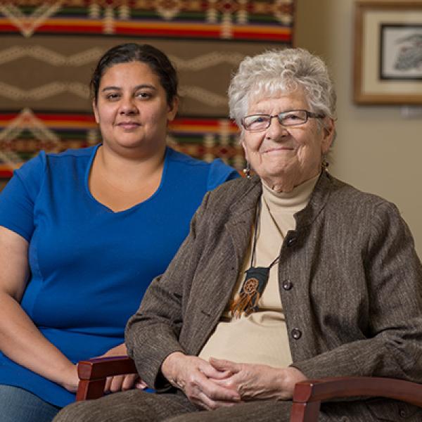 A young woman in blue and an older woman wearing a brown jacket and necklace that looks like a pouch with leather fringe sit side-by-side in a room in front of a patterned wall hanging