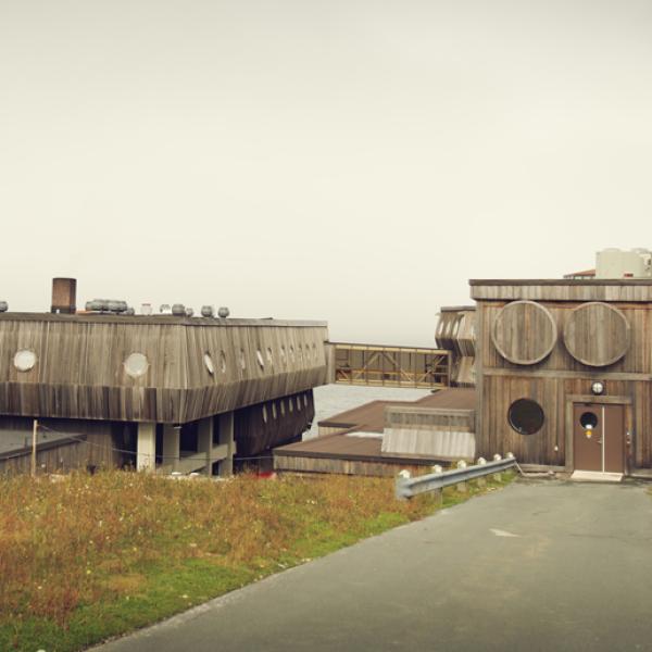 A wooden building resembling a ship with round portholes is perched on a coast line with fog on the opposite shore.