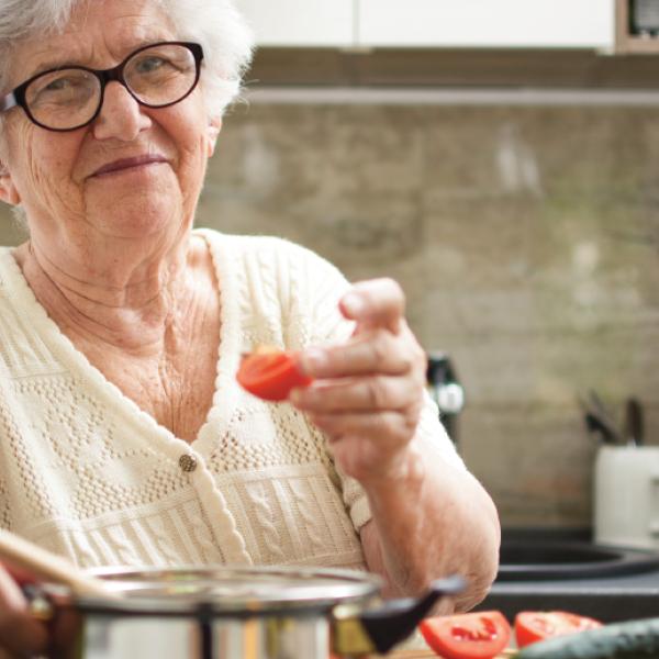 Un quartier de tomate à la main, une femme aux cheveux blancs se tient debout devant un chaudron sur une cuisinière