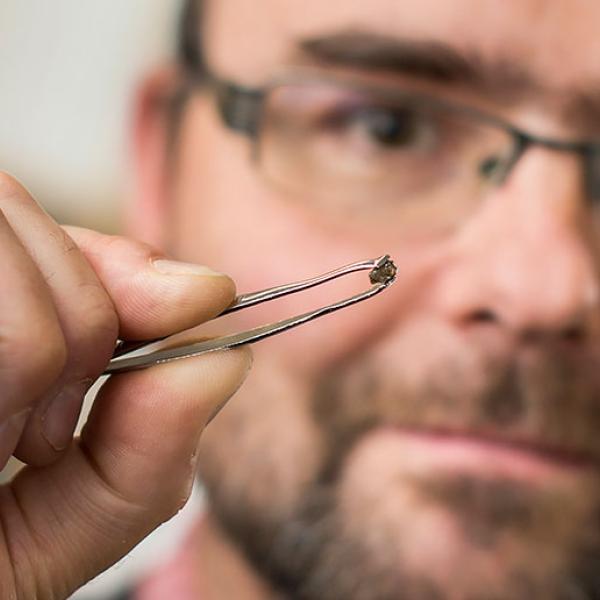 A man uses tweezers to hold up a small diamond. The diamond is in focus, blurring out the man’s face.