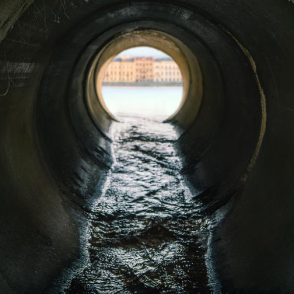 Dark liquid flows through a concrete pipe with a river and cityscape visible through the end of the pipe