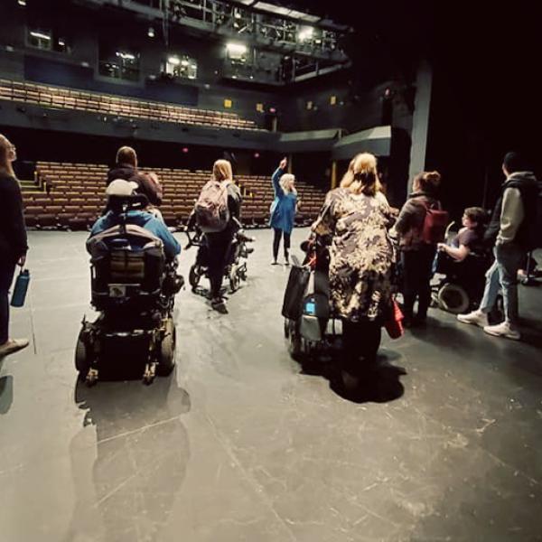 Viewed from backstage, a group of people on a tour stand on stage looking toward the empty auditorium