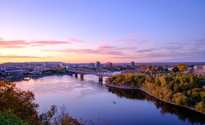 Photo de la rivière des Outaouais vue de la colline du parlement au coucher du soleil. On y voit le musée canadien de l’histoire, la ville qui s’étend, le pont Alexandra qui enjambe la rivière ainsi que de la verdure au premier plan.