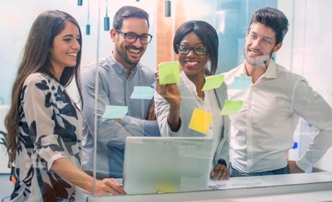 Un groupe de personnes souriantes se trouve devant une cloison en verre couverte de Post-It de couleur bleu, vert et jaune. 