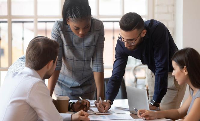 A group of people sitting and standing  around a table look through paperwork and documents