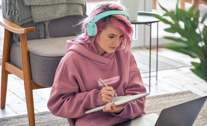 A young person sitting in front of laptop taking notes on a notebook.