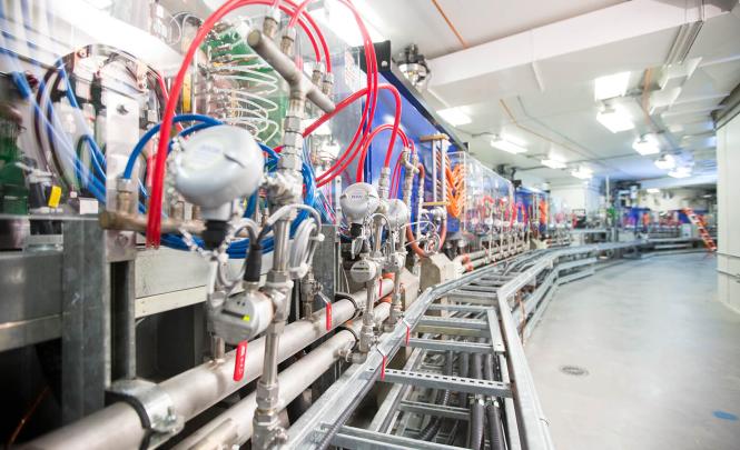 Photo d'un large couloir circulaire dont le sol est en béton. Sur le mur gauche se trouvent des instruments de mesure, des tuyaux en acier et en plastique de diverses couleurs et des valves.