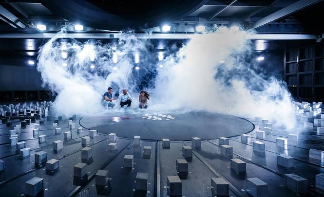 Three people sitting in a test chamber in front of a miniature town, surrounded by gusts of white smoke and wind.