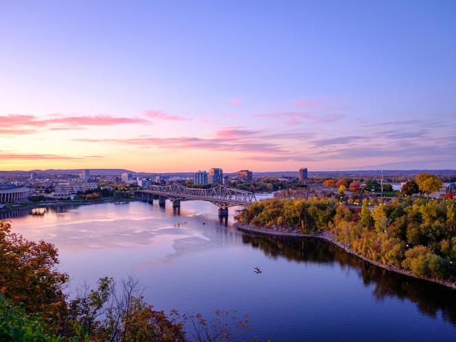 Photo de la rivière des Outaouais vue de la colline du parlement au coucher du soleil. On y voit le musée canadien de l’histoire, la ville qui s’étend, le pont Alexandra qui enjambe la rivière ainsi que de la verdure au premier plan.
