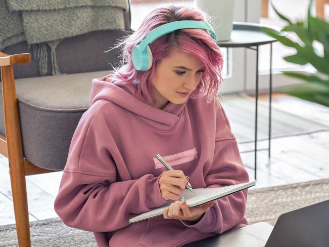 A young person sitting in front of laptop taking notes on a notebook.