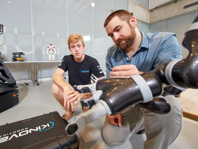 A researcher crouching and demonstrating a robotic arm to an observer.