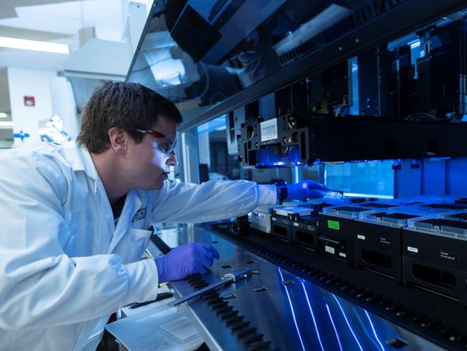 Person wearing a lab coat and gloves loading microplates into an automated liquid-handling system.