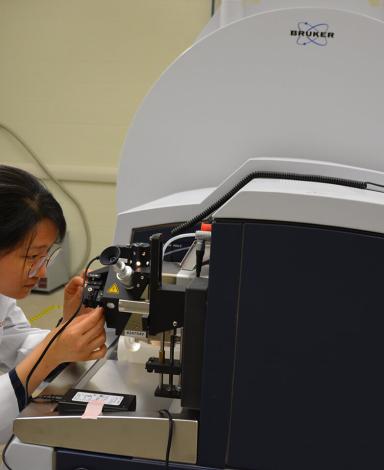 A person in a lab coat looks closely at an apparatus that is part of a black and grey machine in a lab.