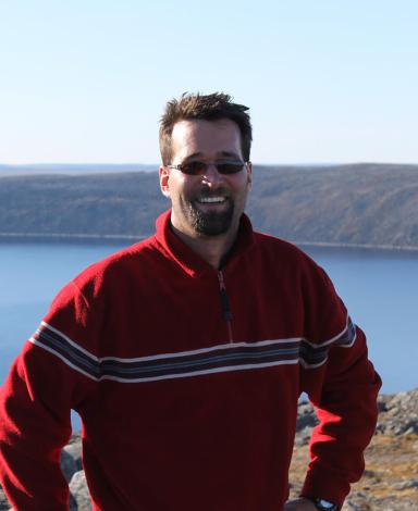 Guillaume St-Onge is wearing a striped red sweater and sunglasses, posing in front of the Pingualuit crater in Quebec, full of clear blue water.